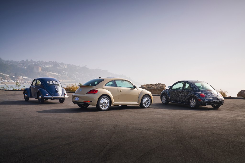 A blue 1949 Volkswagen Beetle, an off-white 2019 Beetle, and a blue printed 1998 New Beetle parked overlooking hills and an ocean