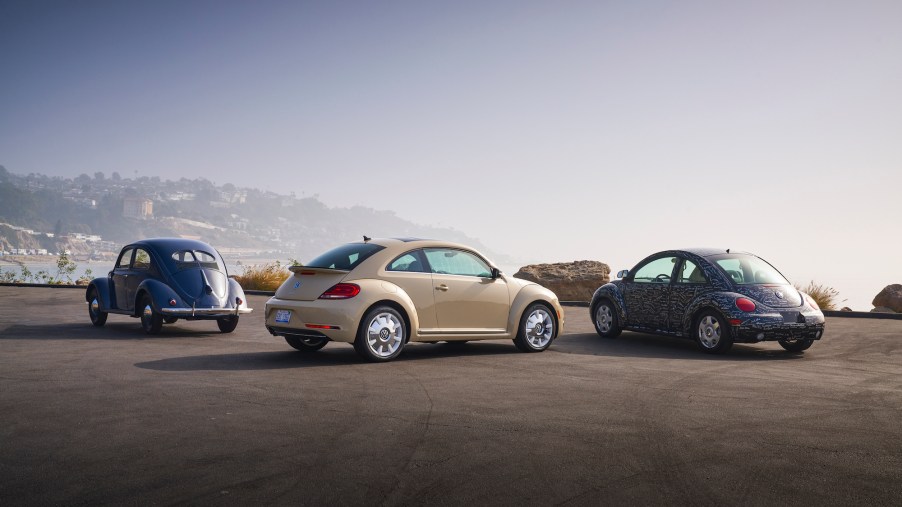 A blue 1949 Volkswagen Beetle, an off-white 2019 Beetle, and a blue printed 1998 New Beetle parked overlooking hills and an ocean