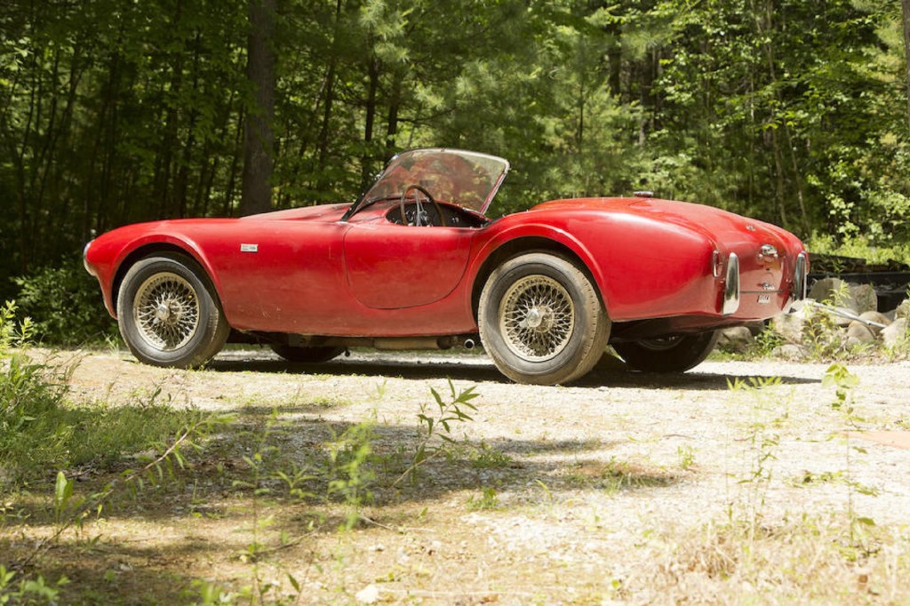 The side 3/4 view of a dusty red 1963 Shelby Cobra 260 parked by a forest