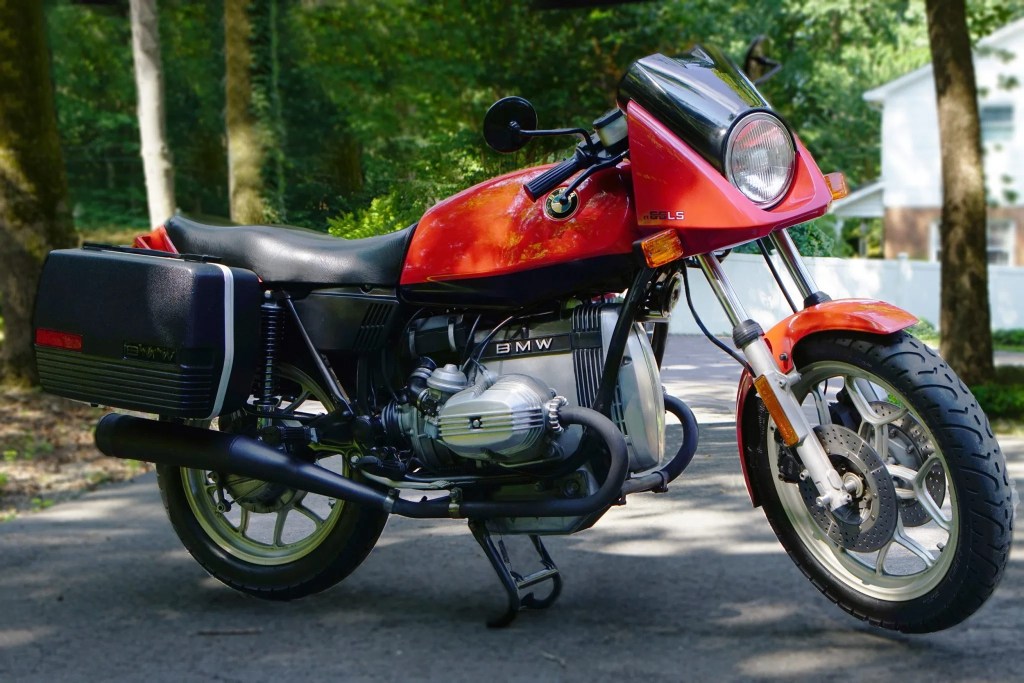 An orange-and-black 1983 BMW R65LS with black panniers on a tree-lined driveway