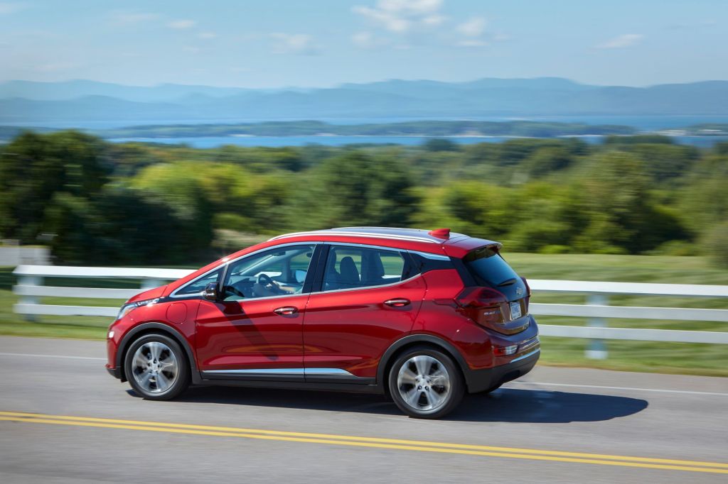 A red 2020 Chevrolet Bolt EV model driving on a highway past a white fence and grass fields