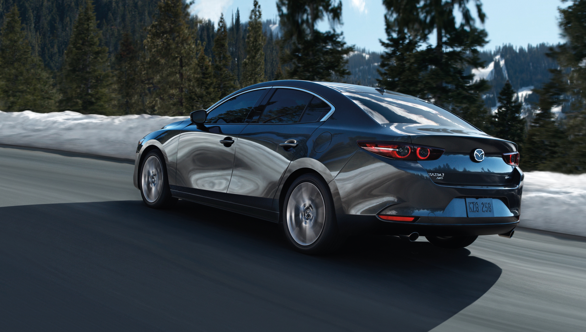 A dark-gray metallic 2020 Mazda3 sedan traveling on a mountain road past snow and pine trees on a sunny day