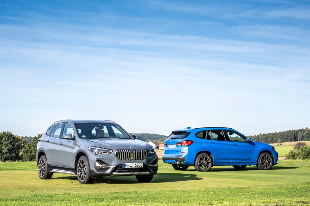 Two 2021 BMW X1 luxury compact SUVs parked on a large green lawn in front of rolling hills and trees
