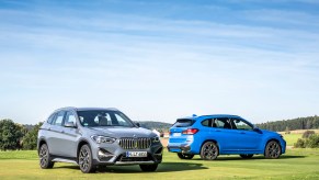 Two 2021 BMW X1 luxury compact SUVs parked on a large green lawn in front of rolling hills and trees