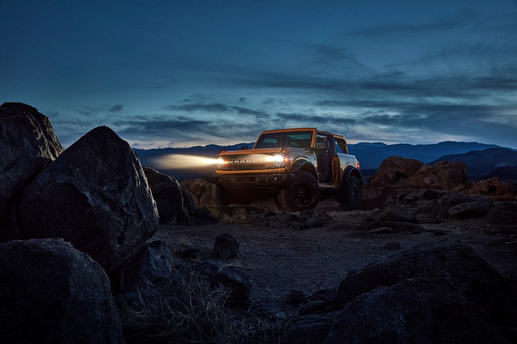 A yellow 2021 Ford Bronco with its headlights on as it sits on rocks in front of mountains at night