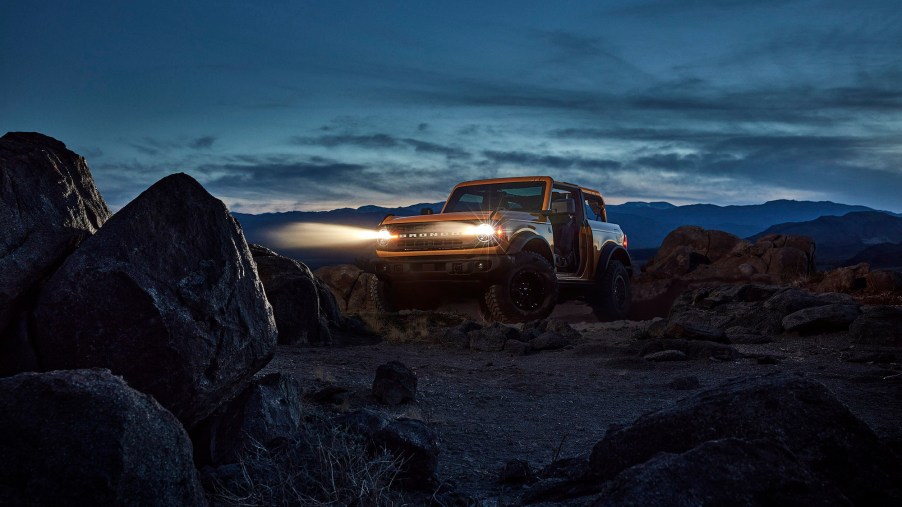 A yellow 2021 Ford Bronco with its headlights on as it sits on rocks in front of mountains at night
