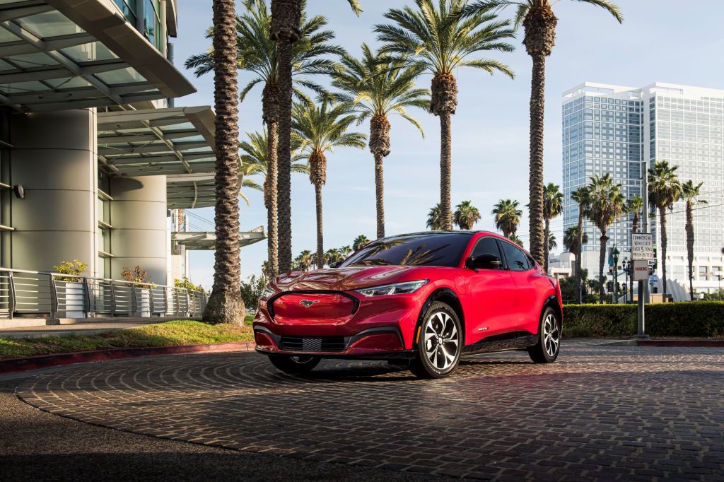 A red 2021 Ford Mustang Mach-E sits on a brick driveway surrounded by palm trees and modern buildings.