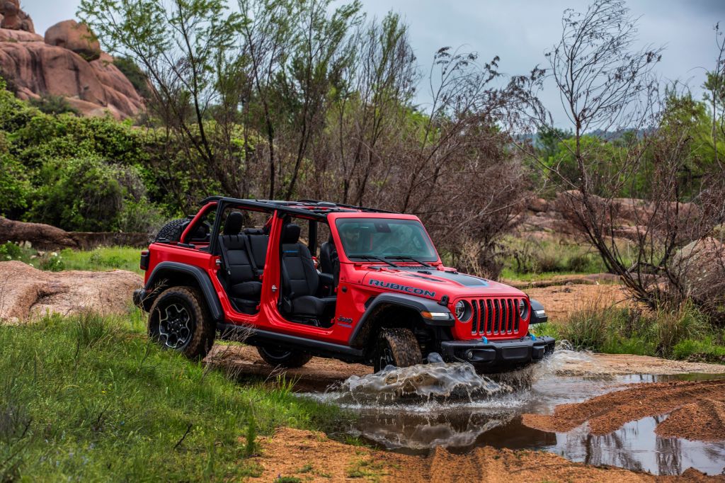 A red 2021 Jeep Wrangler Rubicon 4xe with no doors or roof splashes through a puddle on a desert trail
