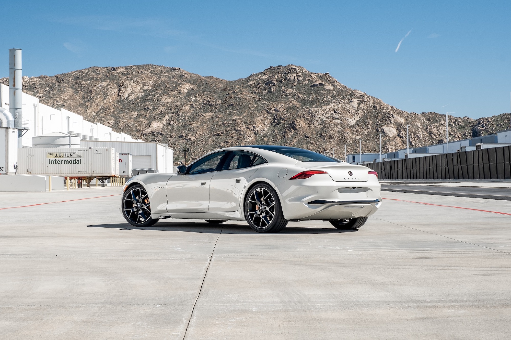 A white 2021 Karma GS-6 sports sedan parked outside a white warehouse in front of a mountain range on a sunny day