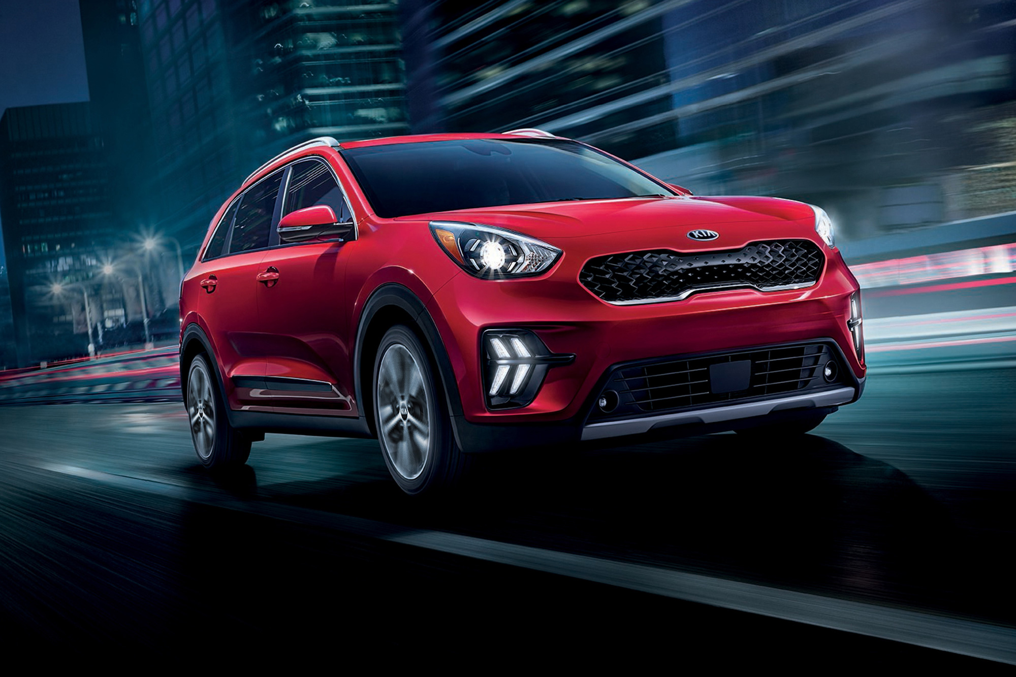 A red 2021 Kia Niro plug-in hybrid compact SUV traveling on a wet city street at night