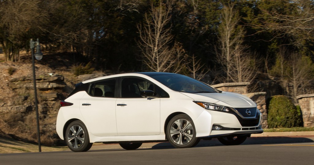 A white 2021 Nissan Nissan Leaf EV parked on a hilly suburban street with trees and a rock wall behind it
