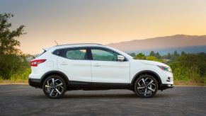 A white side-view of the 2021 Nissan Rogue on a blacktopped area with a background of a clear sunset with hills and trees.