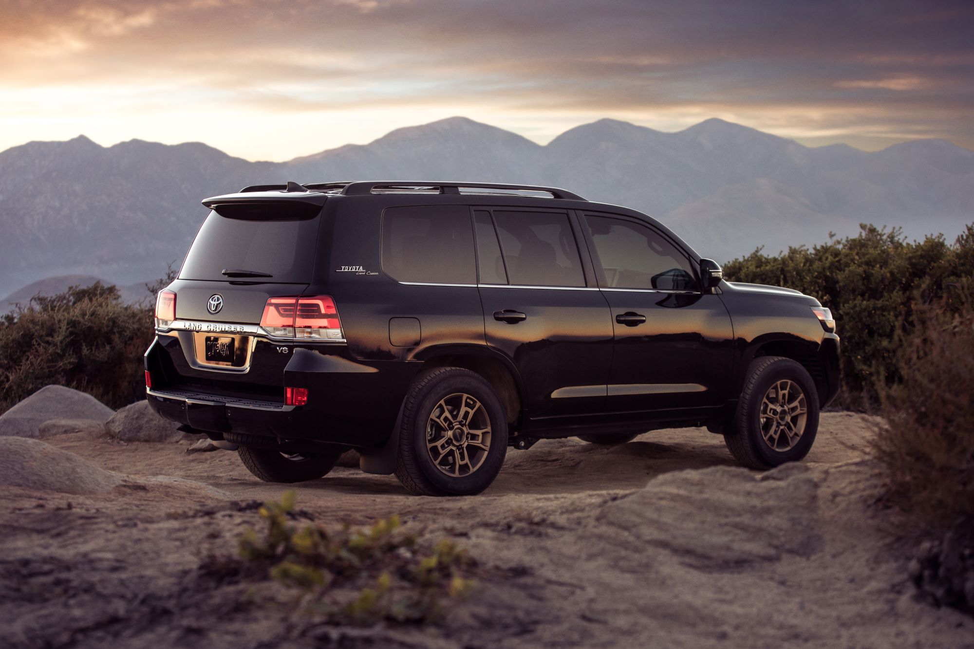 A 2021 Toyota Land Cruiser SUV parked on a rocky plain as the sun sets over a mountain range
