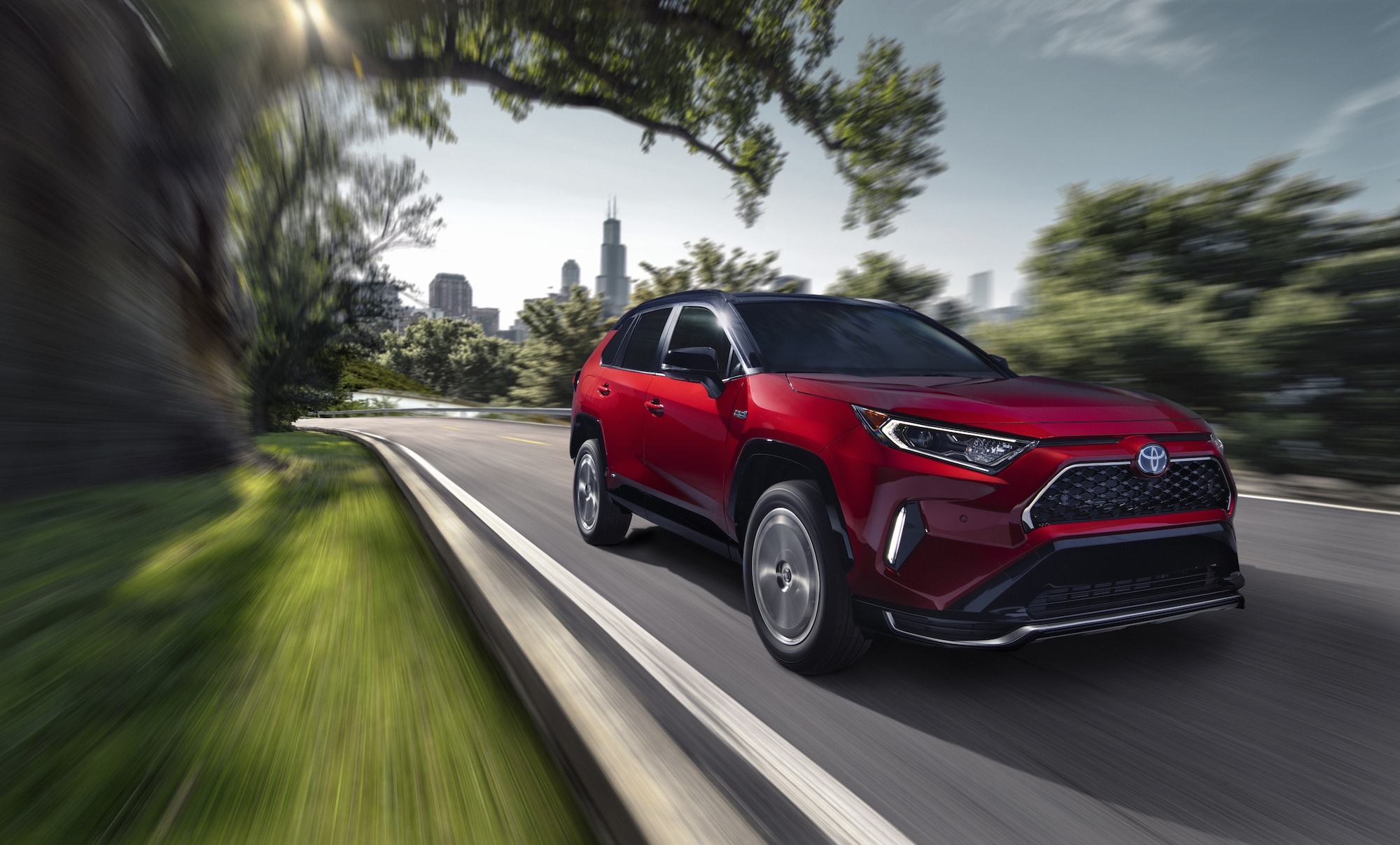 A red 2021 Toyota RAV4 Prime plug-in hybrid compact SUV traveling on a road away from a city