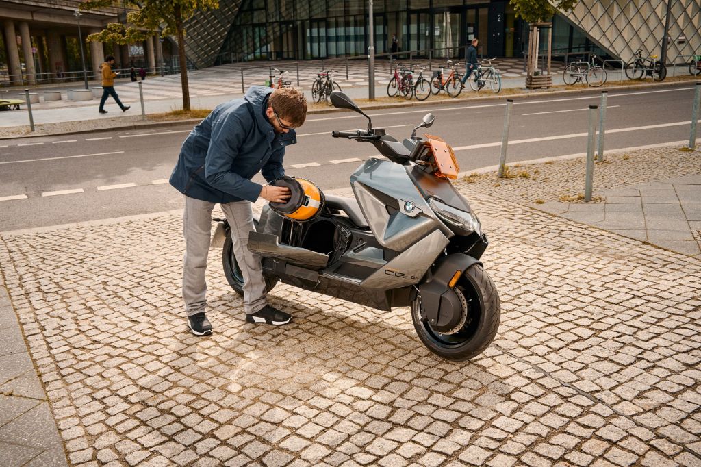 A blue-and-gray-clad rider retrieves their helmet from the under-seat storage compartment of a parked gray-and-orange 2022 BMW CE 04