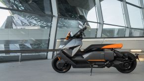The side view of a gray-and-orange 2022 BMW CE 04 by a steel-and-glass building