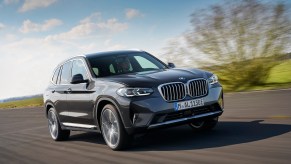 A dark-gray metallic 2022 BMW X3 xDrive30e luxury compact SUV travels on a country road along green hills