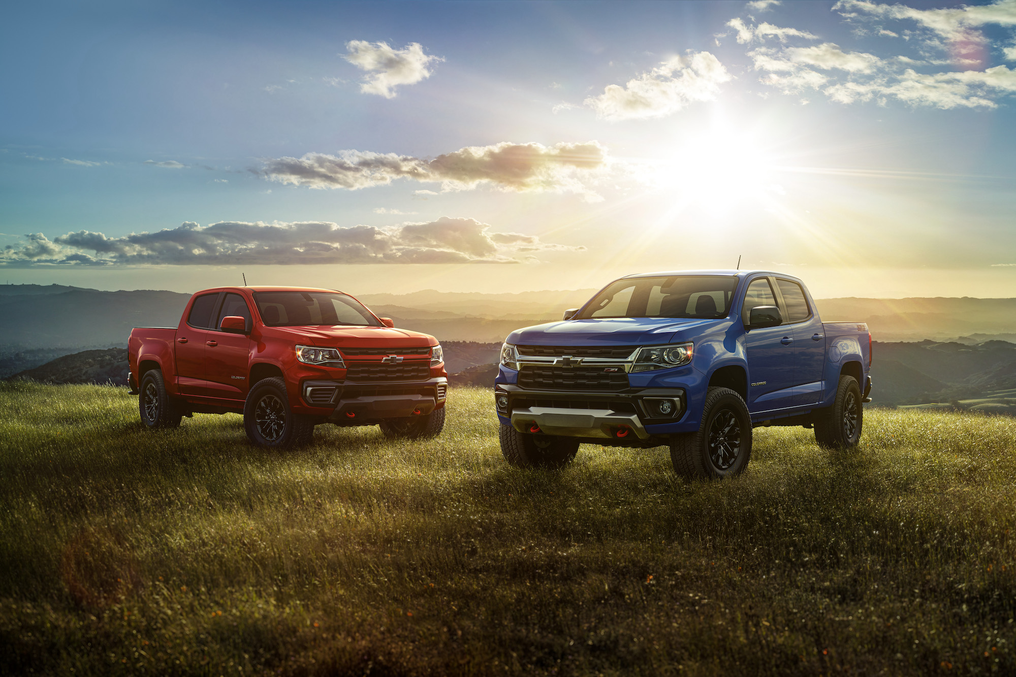 Two 2022 Chevrolet Colorado Trail Boss pickup trucks parked on a grassy hill with the sun and mountains behind them