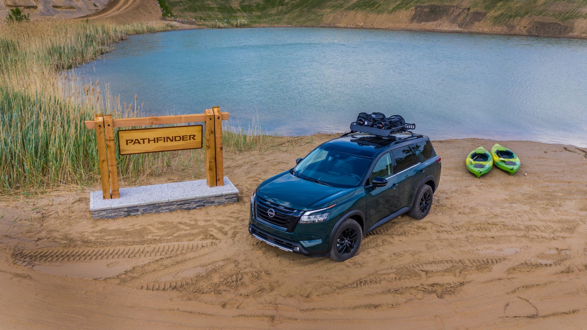 A blue 2022 Nissan Pathfinder sits in front of a pond on a dirt spot between a wooden sign with a concrete base that reads 'Pathfinder' and two kayaks that are light green in color.