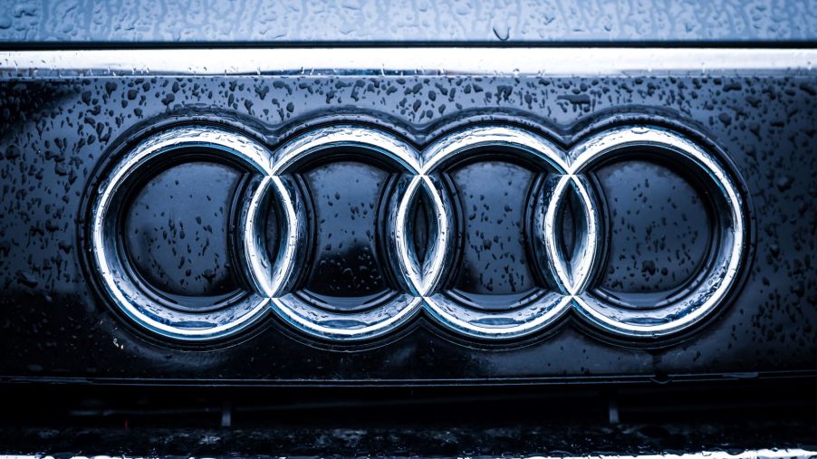 Audi's logo of the four connected rings are placed on a black background of a car grille with raindrops.