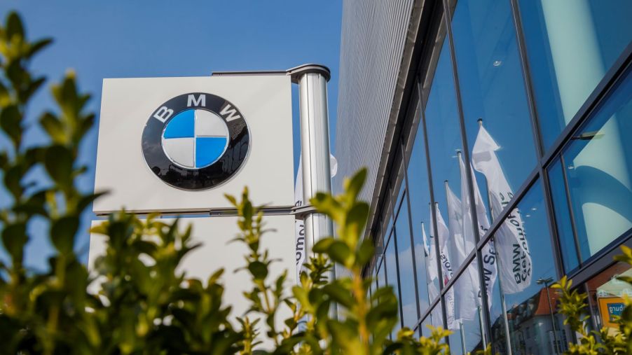 A BMW dealership marque is beside of a building with large windows with some blurred green shrubbery in the front.