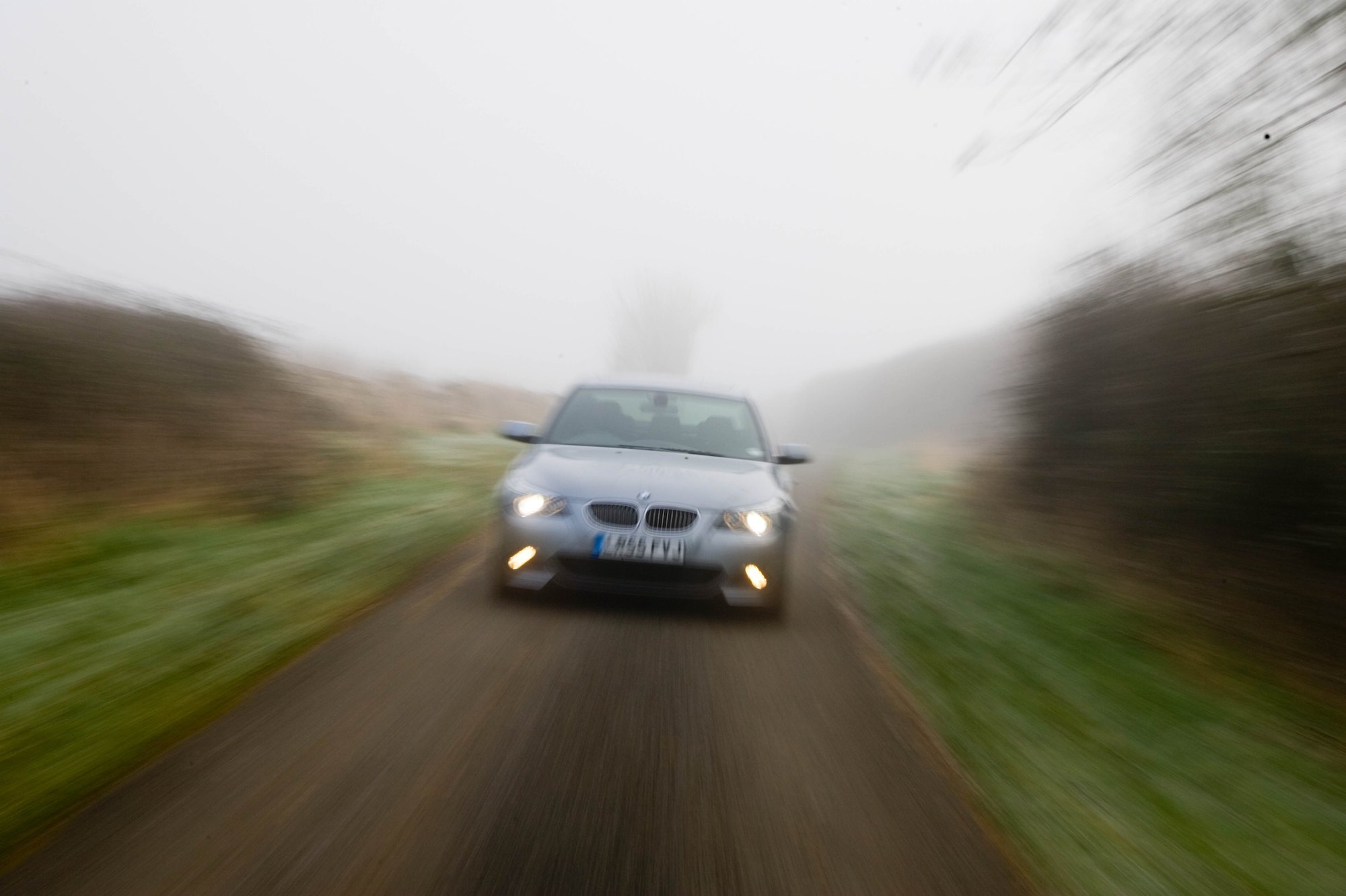 A BMW 5-Series car drives with its headlights illuminated on a country road in foggy weather