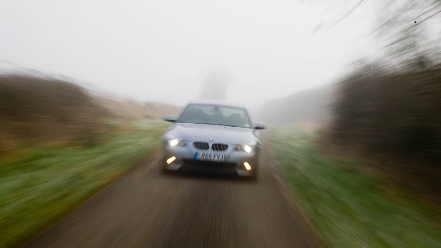 A BMW 5-Series car drives with its headlights illuminated on a country road in foggy weather