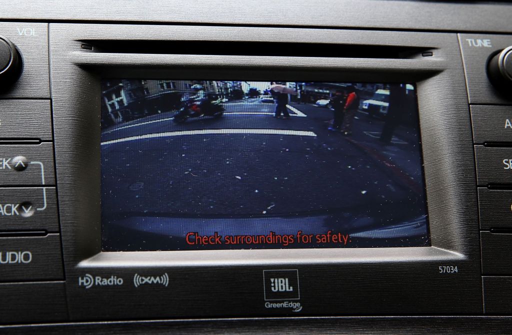 A infotainment screen with a backup camera view that has pedestrians and motorcyclists in the view of the camera with 'Check surroundings for safety' in red at the bottom of the screen.