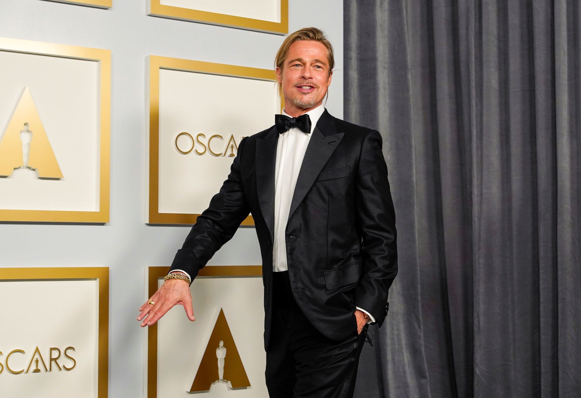 Brad Pitt walking in a black suit, white button up shirt and bow tie in front of a grey backdrop with gold references to the Oscars on it.