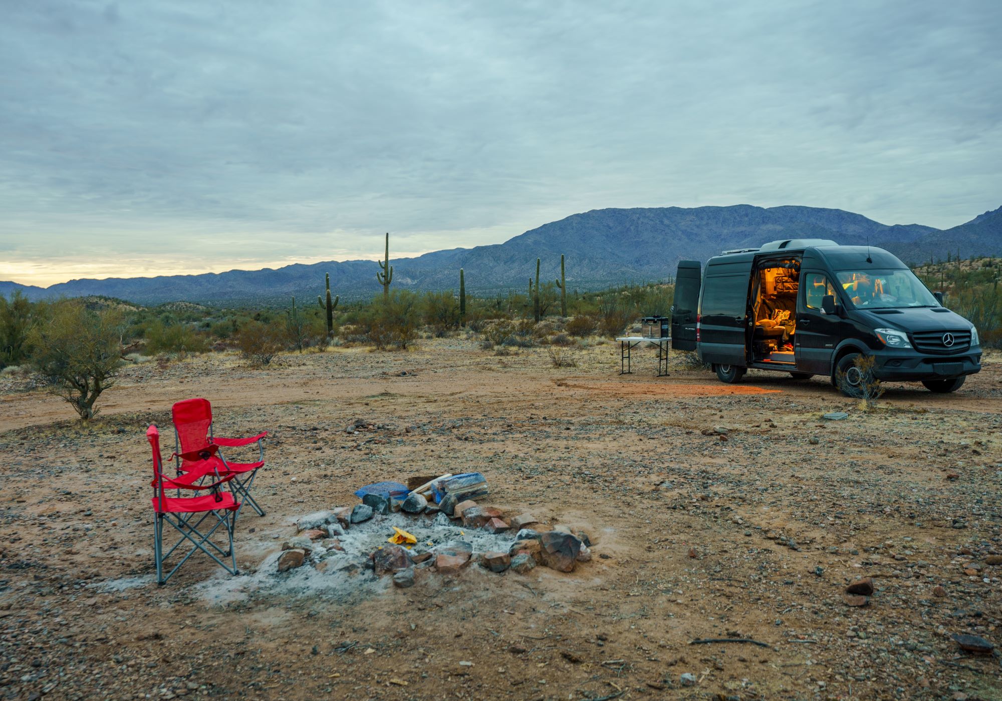 A rental camper van sits in a desert style area with a red chair sitting next to a campfire that recently went out.