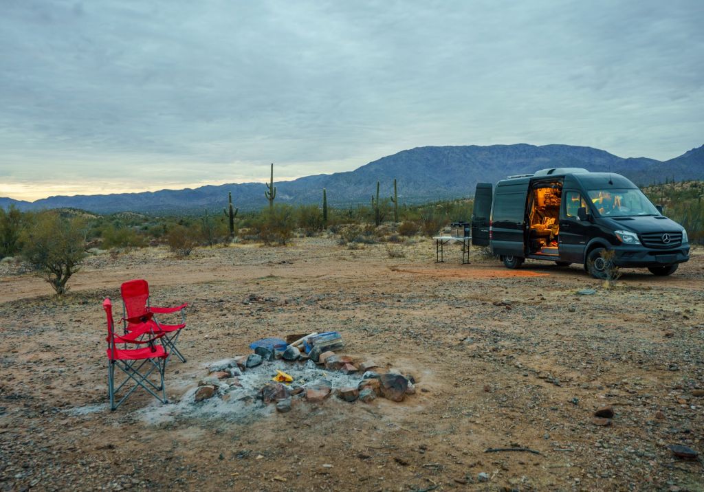 A camper van sits in a desert style area with a red chair sitting next to a campfire that recently went out.