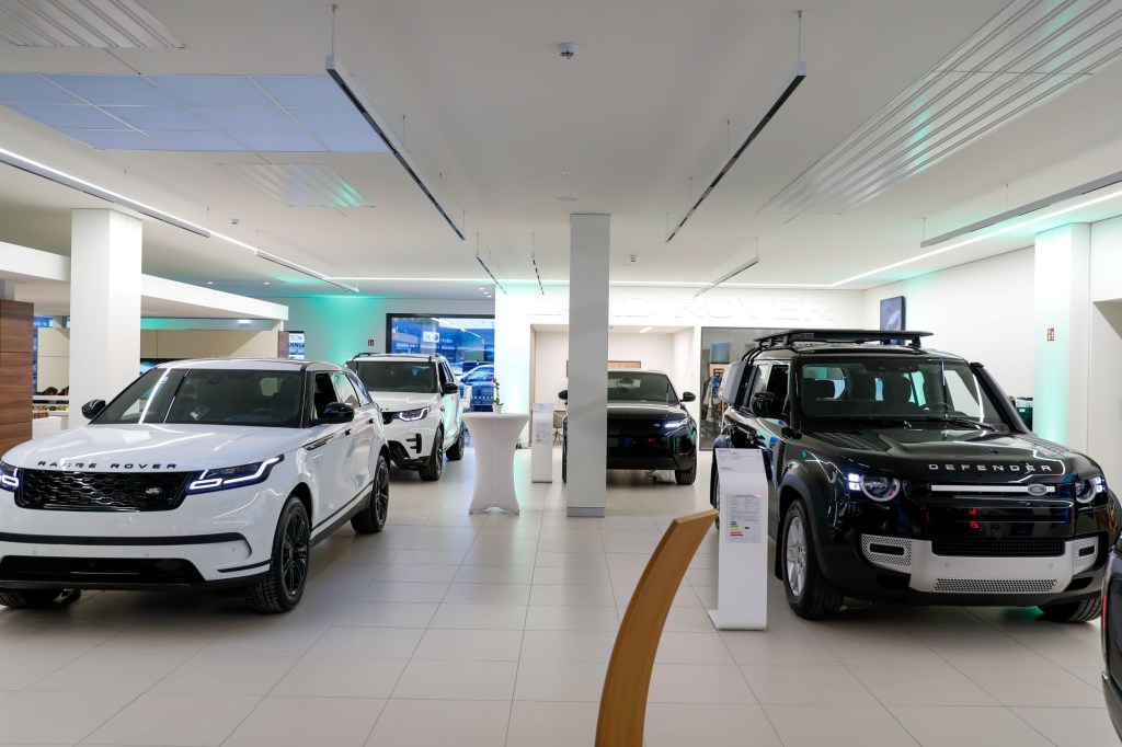 Picture of the inside of a car dealership with four Land Rovers, two on the right that are black and two on the let that are white, against a background of white walls, a wall with screens and a white column in the center.