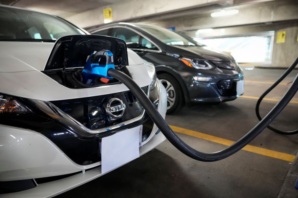 A white Nissan Leaf EV plugged into an electric charging station in a parking garage