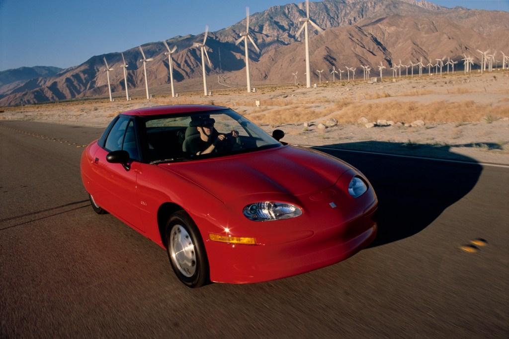 A red GM EV1 parked on a road alongside wind turbines and mountains