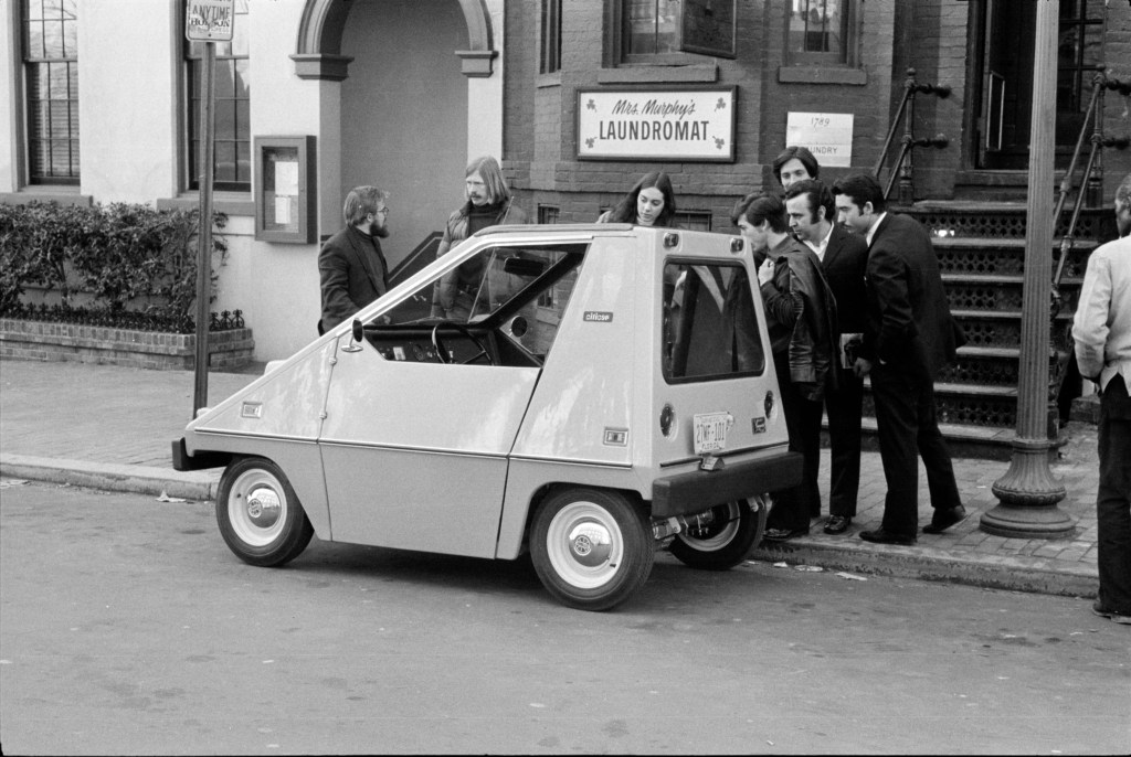 People standing around Sebring-Vanguard CitiCar parked on side of the road.