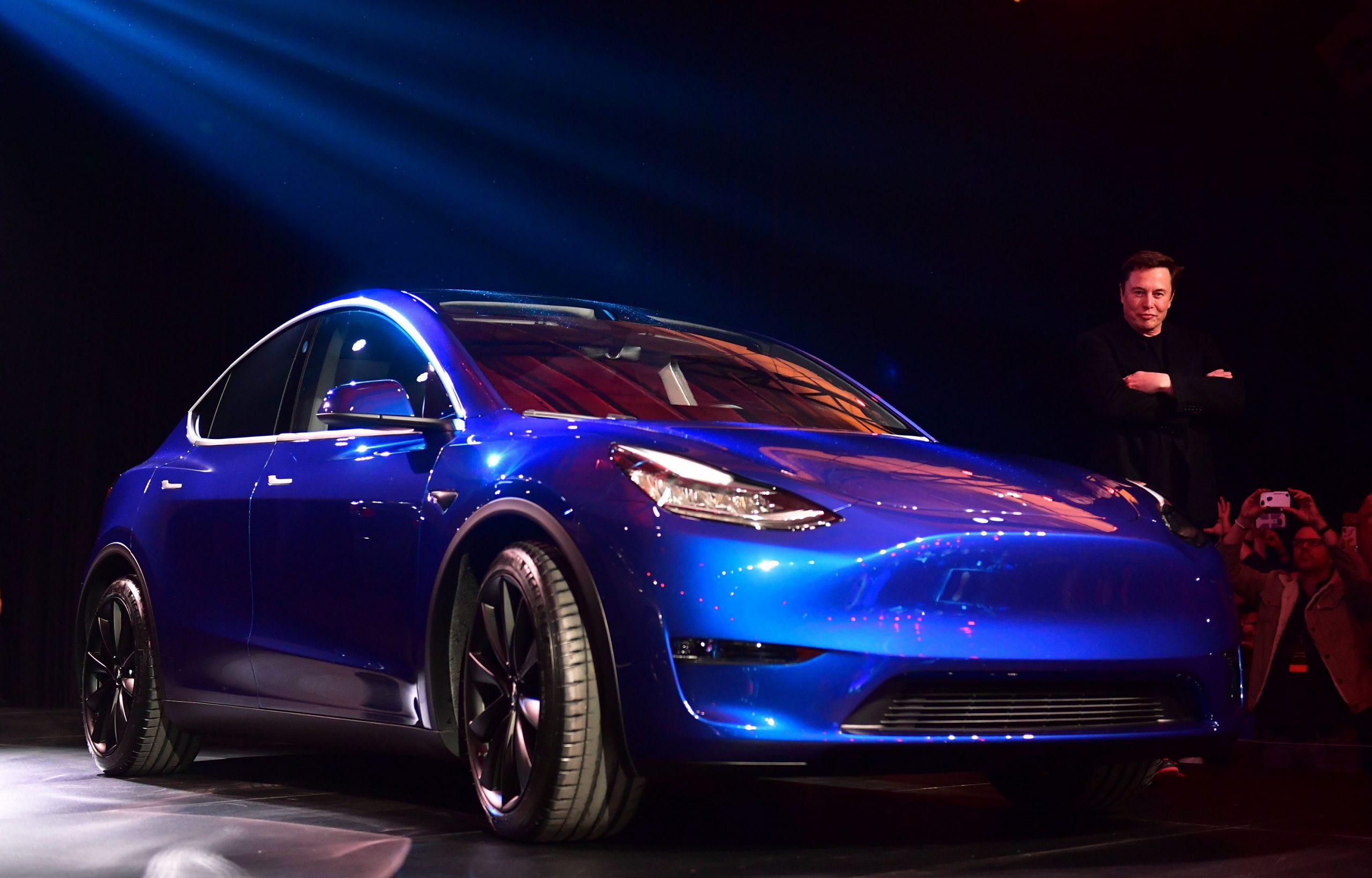 Tesla CEO Elon Musk stands next to a blue Tesla Model Y SUV.