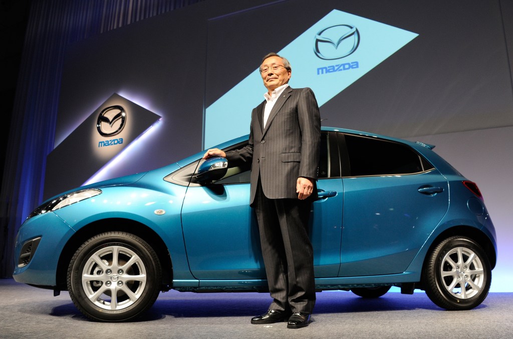 a blue 2011 Mazda Mazda3 hatchback alongside the Mazda president at a press conference. 