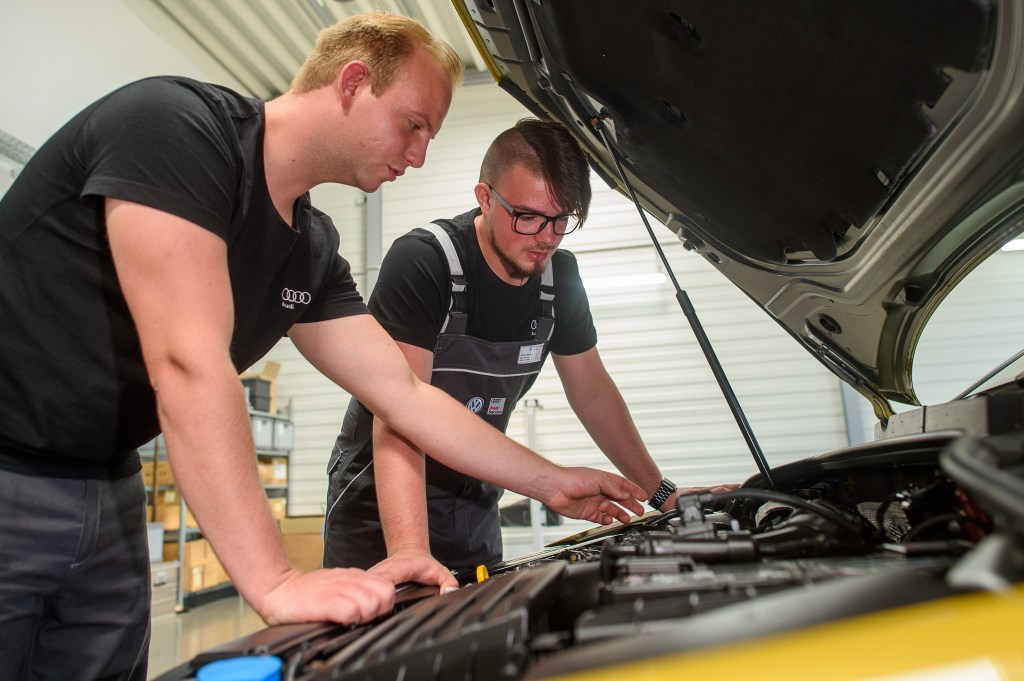 Two mechanics peer into the engine bay of a car in for repairs