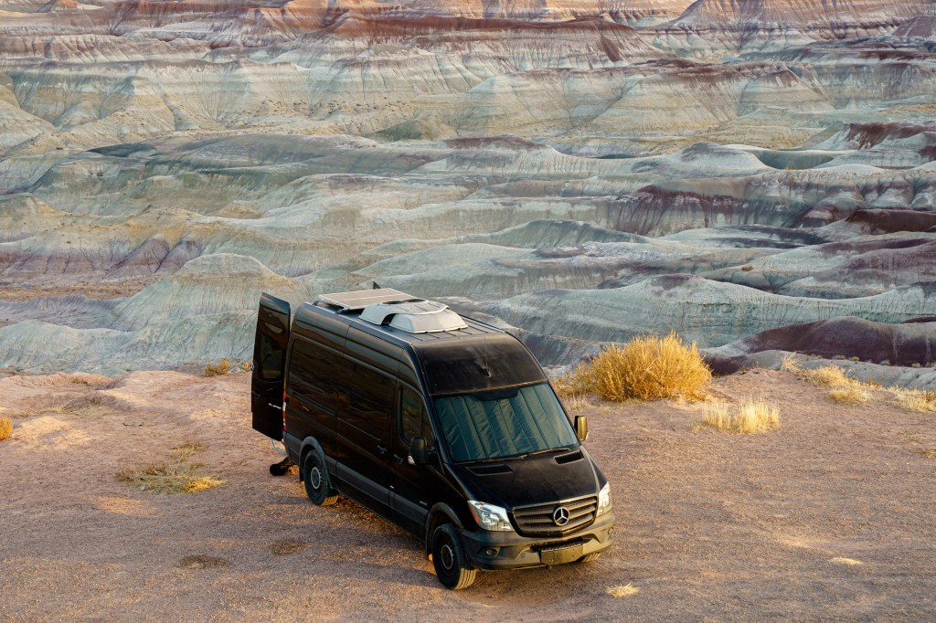 A black Mercedes-Benz Sprinter camper sits in the desert at sunset