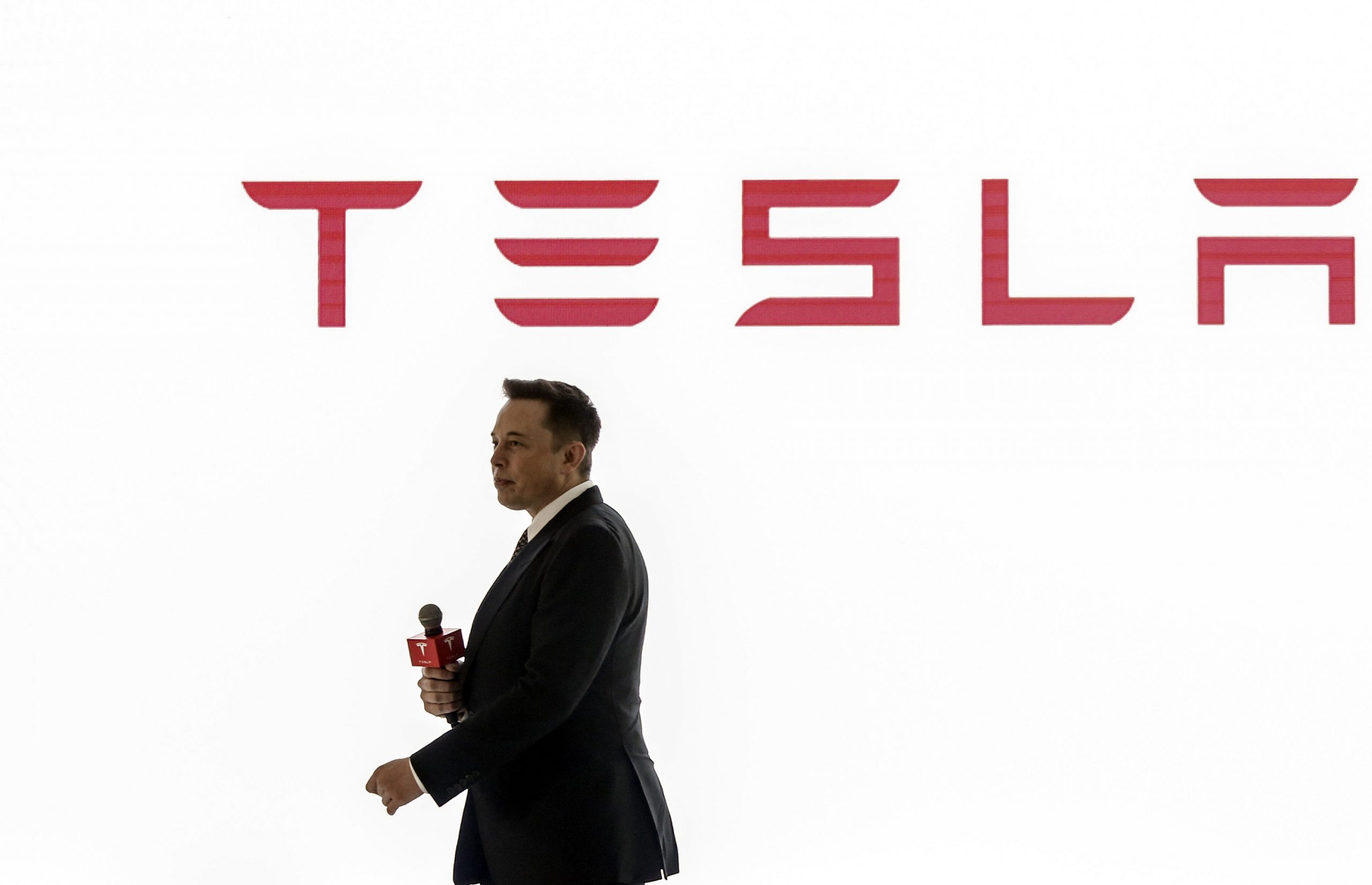 Tesla CEO Elon Musk presents at a conference with the brand's logo seen in red on a white background behind him