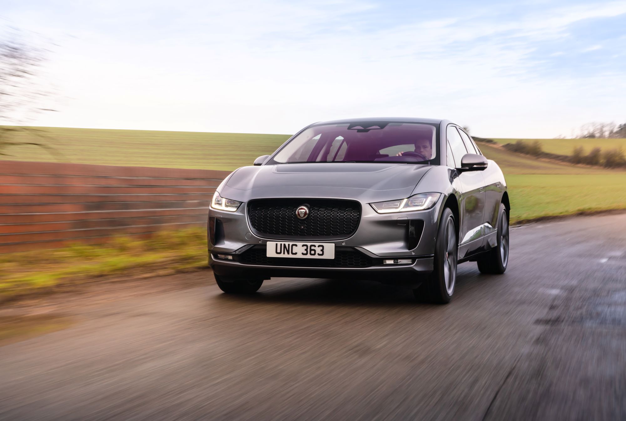 A silver Jaguar I-Pace driving on a highway with a background of green plains.