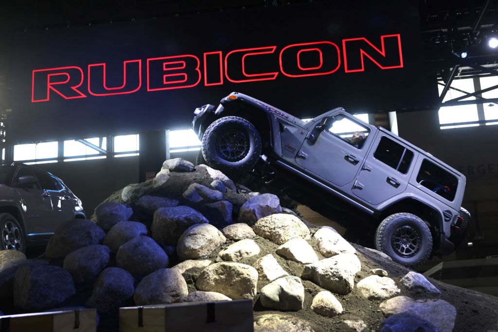 A grey Jeep Rubicon climbing a hill of large rocks with 'Rubicon' printed in red lettering filled in with black in the background.