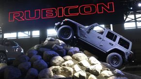 A grey Jeep Rubicon climbing a hill of large rocks with 'Rubicon' printed in red lettering filled in with black in the background.