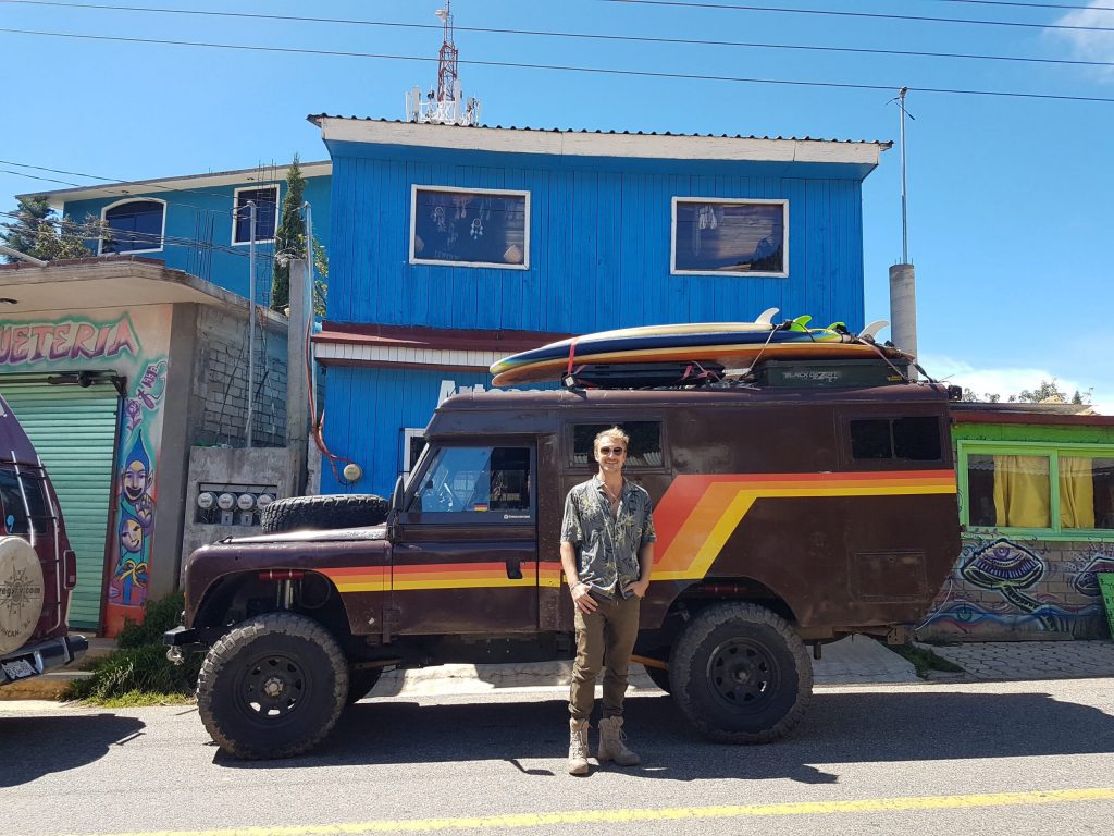 This vintage Land Rover Defender camper is the coolest thing on four wheels. Parked in front of a bright blue building with surfboards on top.