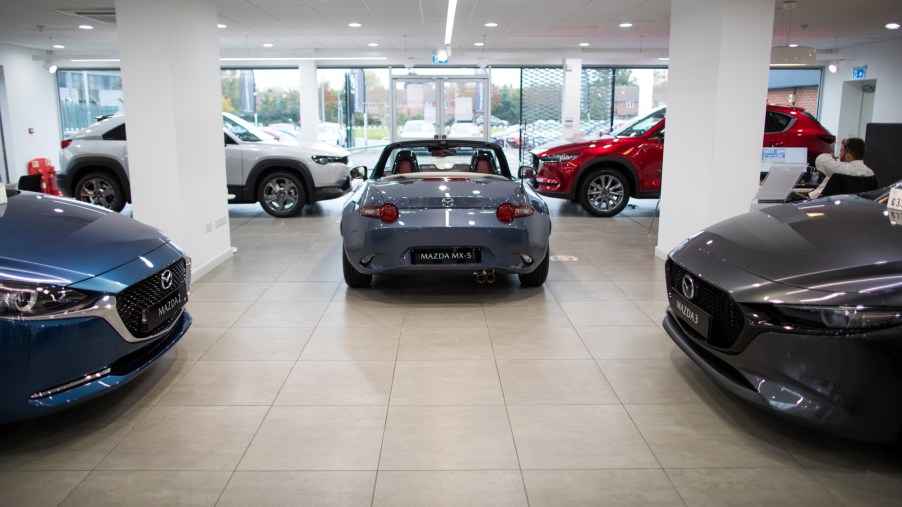 Various Mazda cars sit in a dealership style room with the back end of a grey Mazda MX-5 in the center, the front hood of a Mazda 2 on the right and a Mazda 3 on the left, with two crossover Mazda's in the back partially blocked by white pillars. All in front of a wall length window.