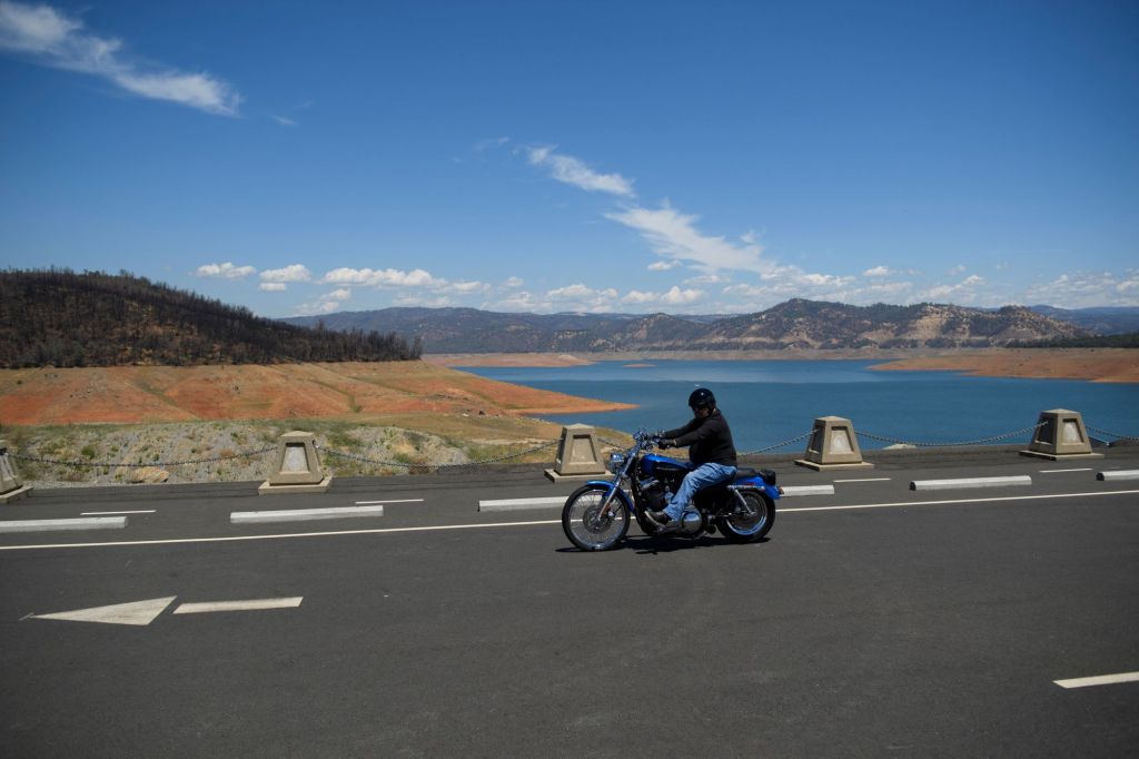 Motorcyclist riding on a black bike dressed in black over a bridge in California with a Mountainous landscape in the background and a brilliant blue body of water.