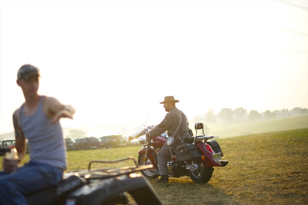 Motorcyclist with a cowboy hat, jeans, denim jacket on a red motorcycle in the middle of field with a bright and foggy sunrise and a few cars blurred in the background.
