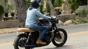 A man with a light blue jean jacket, dark blue jeans, brown boots, and a black helmet riding on a highway on a black triumph motorcycle with a tan seat. The background is blurred with palm trees and other forms of tree trunks and a front section of an adobe style home.