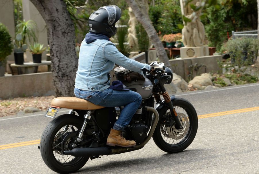 A man with a light blue jean jacket, dark blue jeans, brown boots, and a black helmet riding on a highway on a black triumph motorcycle with a tan seat. The background is blurred with palm trees and other forms of tree trunks and a front section of an adobe style home.