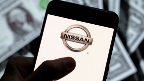 Photo illustration of the Nissan logo displayed on a smartphone screen with U.S. dollar bills in the background